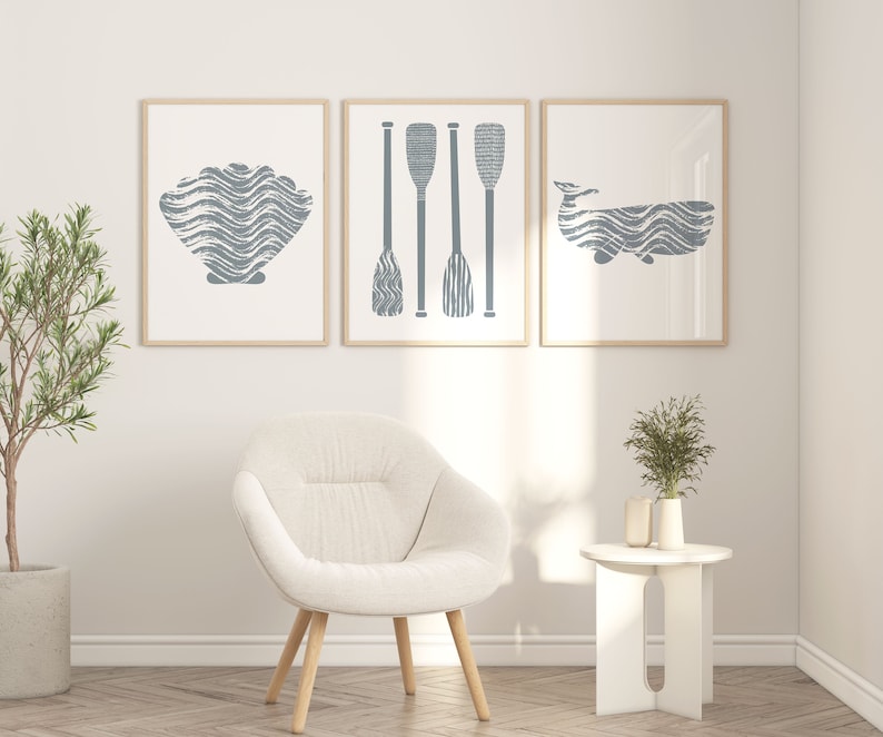Puede incluir: Tres impresiones enmarcadas con un tema n&aacute;utico. Las impresiones presentan una concha, tres remos y una ballena. Las impresiones est&aacute;n colgadas en una pared blanca sobre un sill&oacute;n blanco y una mesa auxiliar blanca.