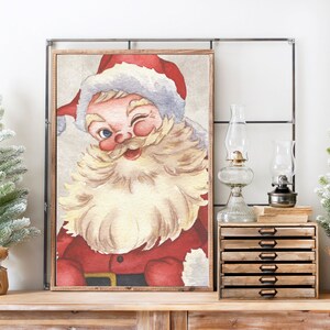 May include: A vintage-style watercolor painting of Santa Claus with a red suit, white beard, and a mischievous wink. The painting is framed in a wooden frame and has a white background.