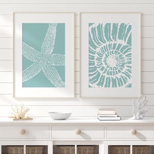 Puede incluir: Dos láminas enmarcadas con motivos marinos. Una muestra una estrella de mar blanca sobre fondo turquesa, la otra una concha de nautilus blanca sobre turquesa. Ambas en marcos de madera clara, ideales para decoración costera.