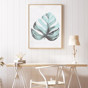 May include: A framed print of a light blue watercolor leaf on a white background. The print is hanging on a white wall above a wooden desk with a white chair and a lamp.