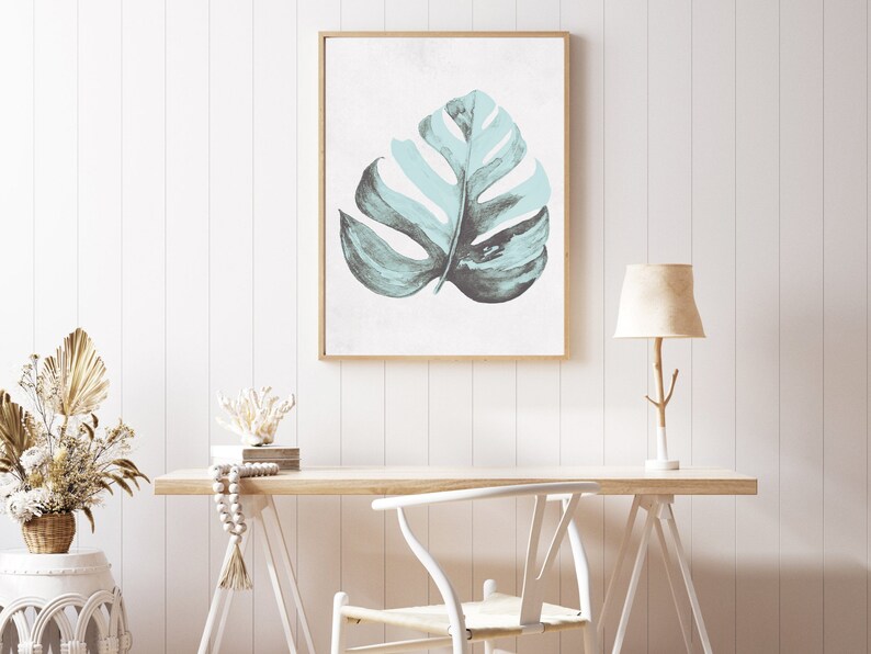 May include: A framed print of a light blue watercolor leaf on a white background. The print is hanging on a white wall above a wooden desk with a white chair and a lamp.