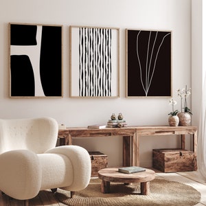 May include: Three black and white abstract art prints with wooden frames hanging on a white wall above a wooden table with a white armchair and a round wooden coffee table.