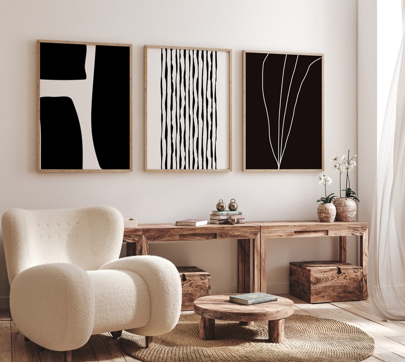 May include: Three black and white abstract art prints with wooden frames hanging on a white wall above a wooden table with a white armchair and a round wooden coffee table.
