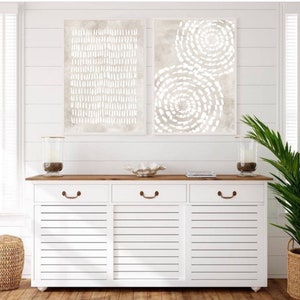 Puede incluir: Dos impresiones de arte abstracto en un esquema de color beige con dise&ntilde;os geom&eacute;tricos blancos. Las impresiones est&aacute;n colgadas en una pared blanca de tablones de madera sobre una c&oacute;moda blanca con tres cajones y una tapa de madera.