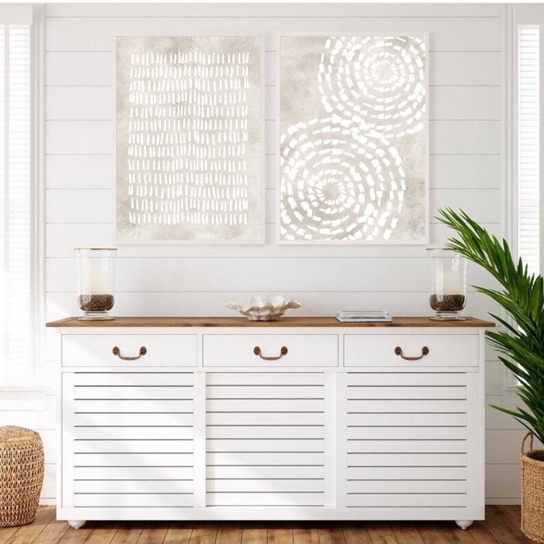 Puede incluir: Dos impresiones de arte abstracto en un esquema de color beige con dise&ntilde;os geom&eacute;tricos blancos. Las impresiones est&aacute;n colgadas en una pared blanca de tablones de madera sobre una c&oacute;moda blanca con tres cajones y una tapa de madera.