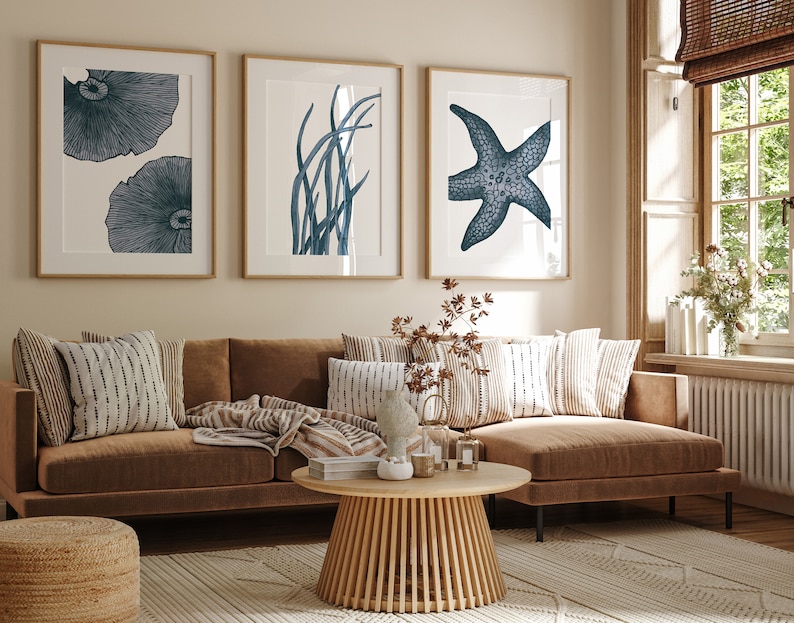 Op de afbeelding: Een bruine hoekbank met kussens en een deken. Drie ingelijste prints met blauwe en witte illustraties van een zeester, zeewier en paddenstoelen hangen aan de muur achter de bank.