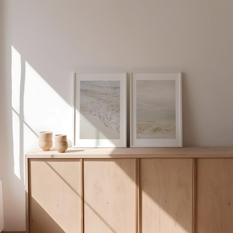 May include: Two framed photographs of seascapes hang above a light wood cabinet with three drawers. The photographs are in white frames and feature a beach scene with waves and a cloudy sky.