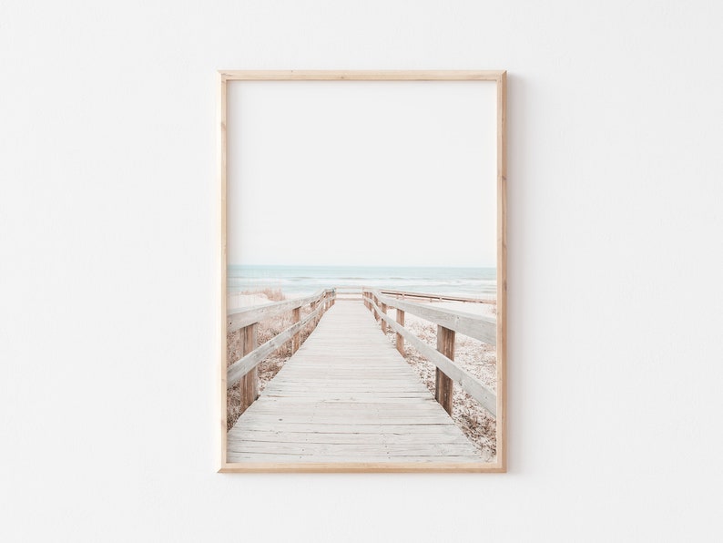 May include: A wooden walkway with railings leads to the ocean. The beach is sandy and the water is a light blue. The sky is a pale blue.