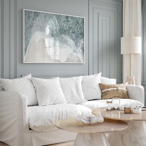 May include: A white sofa with white cushions and a brown throw pillow. There are two round coffee tables in front of the sofa, one with a white shell on top. The sofa is in front of a grey wall with a framed print of an aerial view of ocean waves.