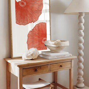 May include: A framed print with two large, abstract, coral-colored shapes on a white background. The print is hanging on a white wall above a wooden table with two drawers. A white ceramic bowl and a book titled "Vincent van Gogh" are on the table.