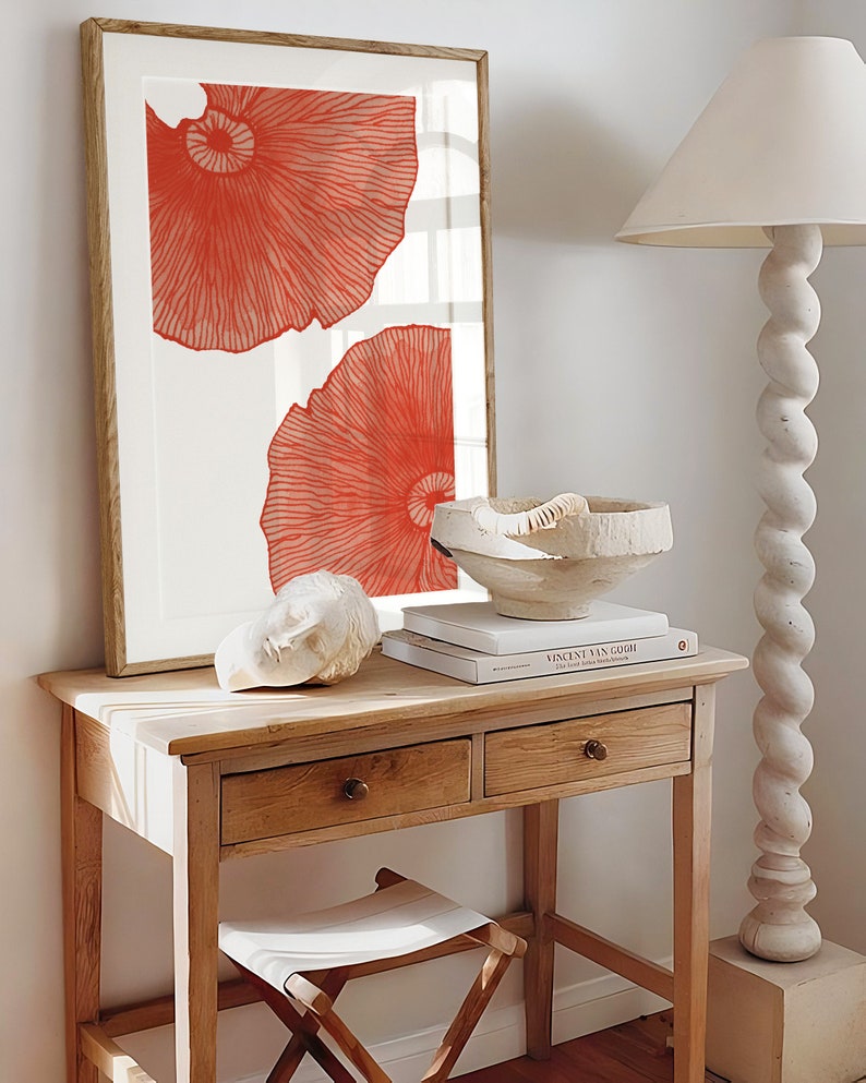 May include: A framed print with two large, abstract, coral-colored shapes on a white background. The print is hanging on a white wall above a wooden table with two drawers. A white ceramic bowl and a book titled "Vincent van Gogh" are on the table.