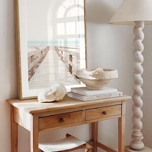 May include: A wooden table with two drawers and a white stool. A framed print of a beach scene with a wooden walkway leading to the sea is hanging on the wall above the table. A white ceramic bowl and a book titled "Vincent van Gogh" are on the table.