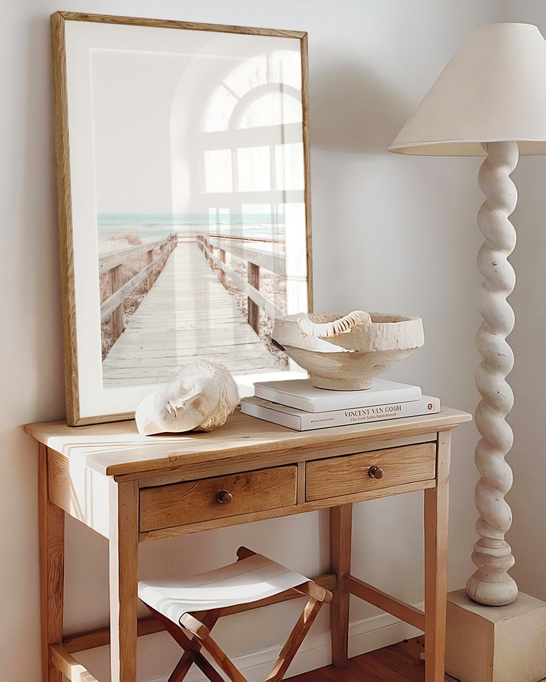 May include: A wooden table with two drawers and a white stool. A framed print of a beach scene with a wooden walkway leading to the sea is hanging on the wall above the table. A white ceramic bowl and a book titled "Vincent van Gogh" are on the table.