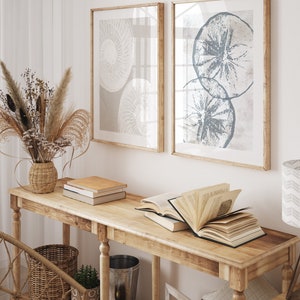 May include: A wooden console table with a light wood finish. The table has two shelves and is decorated with a vase of dried flowers, books, and a decorative bowl. Two framed prints with abstract designs hang on the wall behind the table.