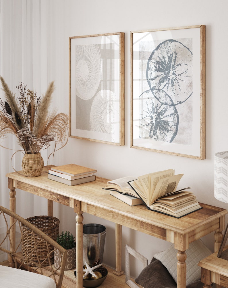 May include: A wooden console table with a light wood finish. The table has two shelves and is decorated with a vase of dried flowers, books, and a decorative bowl. Two framed prints with abstract designs hang on the wall behind the table.