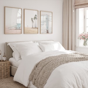 May include: A white bed with a beige knitted throw blanket and three framed prints of beach scenes above the headboard. The prints feature a lifeguard stand, palm trees, and a surfboard.