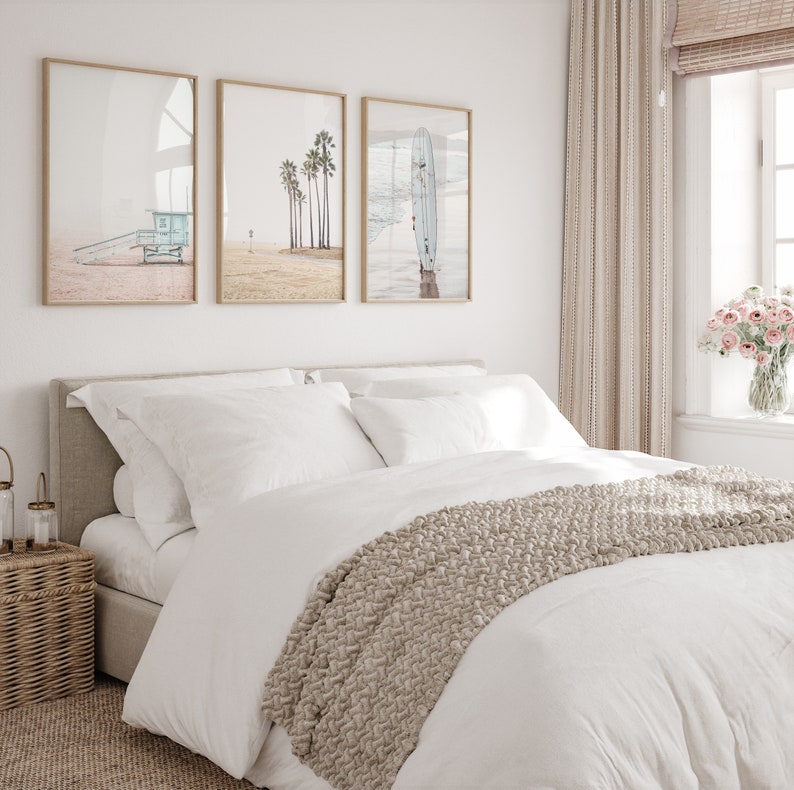 May include: A white bed with a beige knitted throw blanket and three framed prints of beach scenes above the headboard. The prints feature a lifeguard stand, palm trees, and a surfboard.