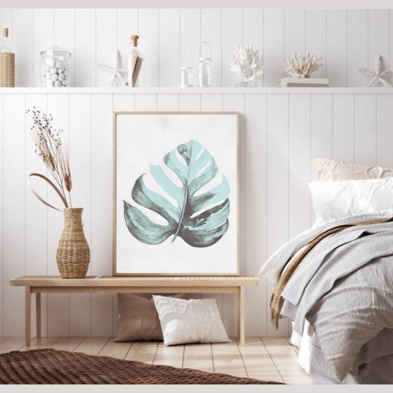 May include: A framed print of a single, large, light blue and dark green tropical leaf on a white background. The print is hanging on a white wall above a wooden bench.