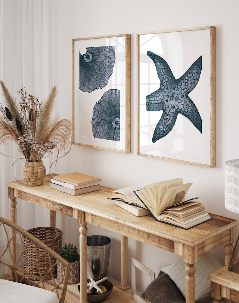Op de afbeelding: Twee ingelijste prints met een witte achtergrond en blauwe illustraties. De print aan de linkerkant toont twee gestileerde blauwe kwallen. De print aan de rechterkant toont een gestileerde blauwe zeester. De prints hangen aan een witte muur boven een houten consoletafel.