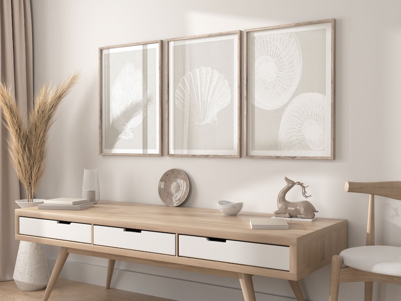 K&ouml;nnte beinhalten: Drei gerahmte Drucke mit beigefarbenem Hintergrund und wei&szlig;en Illustrationen von Muscheln. Die Drucke h&auml;ngen an einer wei&szlig;en Wand &uuml;ber einem hellen Holz-Konsolentisch mit drei wei&szlig;en Schubladen. Eine kleine Hirschfigur und eine Schale stehen auf dem Tisch.