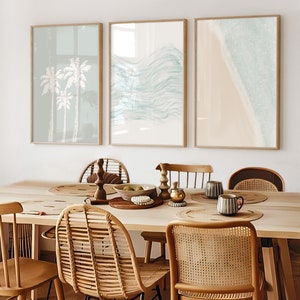 May include: Three framed prints with a beach theme. The first print features palm trees, the second print features abstract waves, and the third print features a beach with waves crashing on the shore. The prints are hanging on a white wall above a wooden table with wicker chairs.
