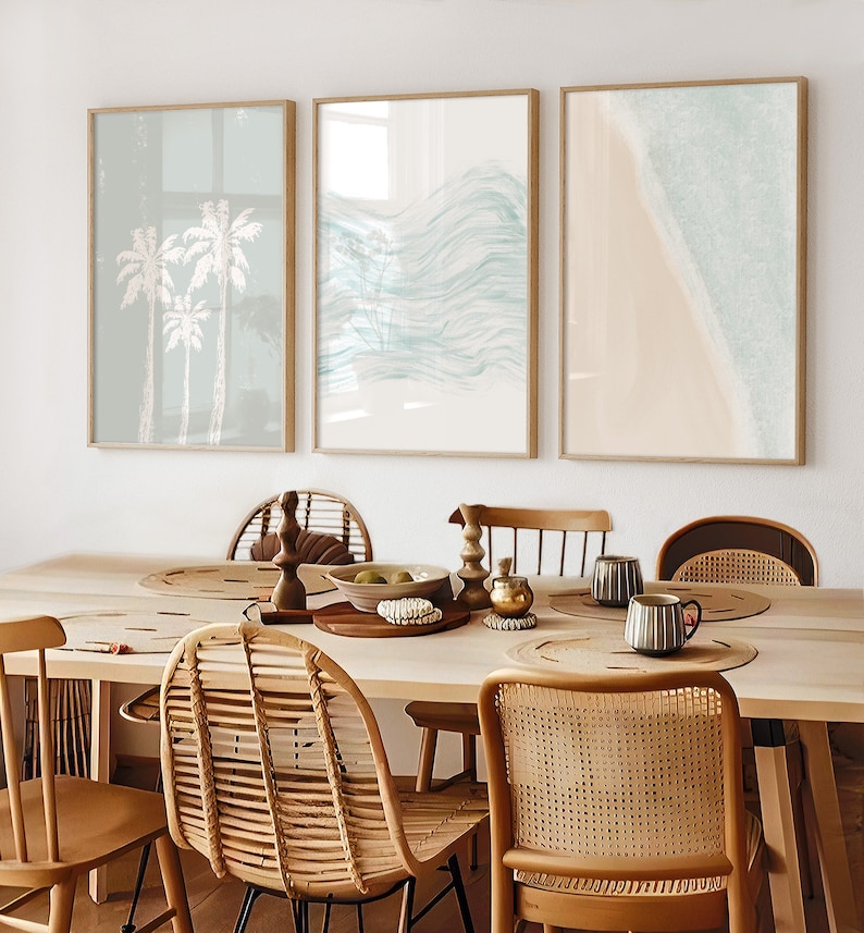 May include: Three framed prints with a beach theme. The first print features palm trees, the second print features abstract waves, and the third print features a beach with waves crashing on the shore. The prints are hanging on a white wall above a wooden table with wicker chairs.