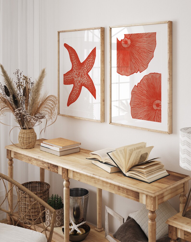 May include: Two framed prints with red illustrations of a starfish and two seashells on a white background. The prints are hanging on a white wall above a wooden console table with books and a vase of dried flowers.