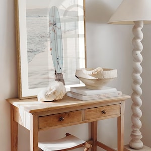 May include: A wooden table with two drawers and a white stool. A framed print of a surfboard in the ocean is on the wall above the table. A white ceramic bowl and a white sculpture are on the table. A book titled "Vincent van Gogh" is on the table.