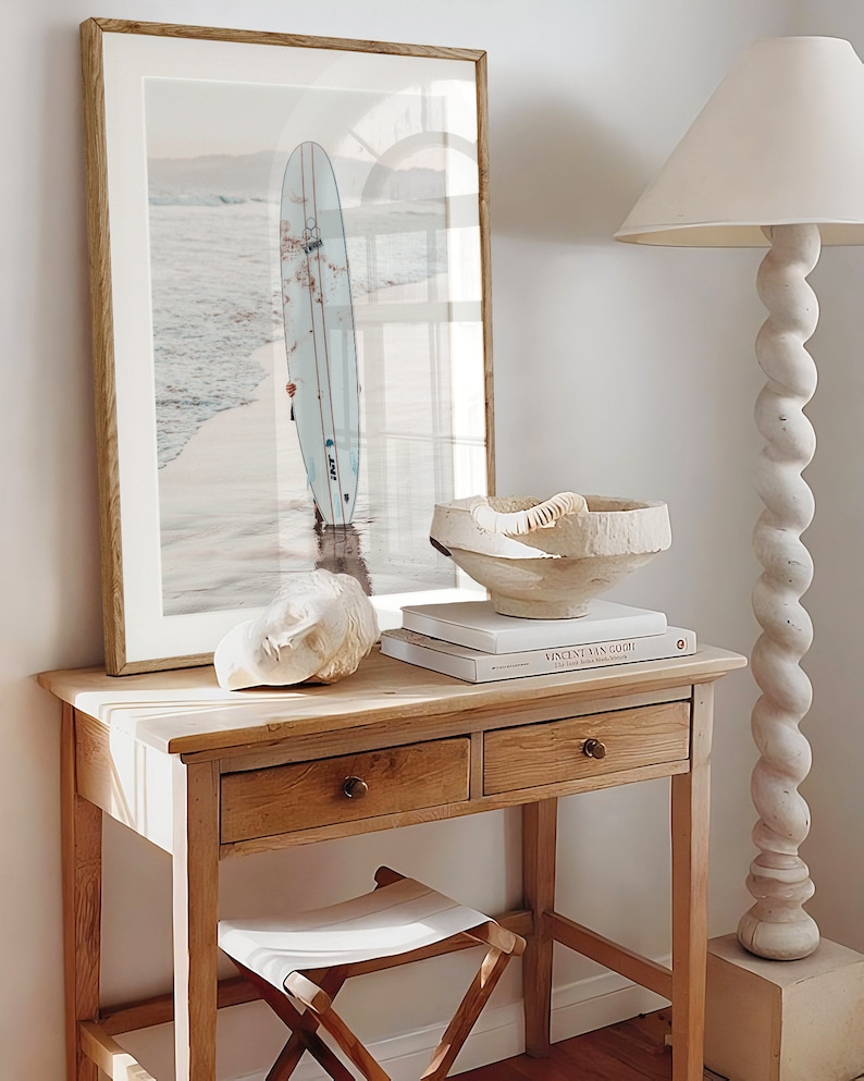 May include: A wooden table with two drawers and a white stool. A framed print of a surfboard in the ocean is on the wall above the table. A white ceramic bowl and a white sculpture are on the table. A book titled "Vincent van Gogh" is on the table.