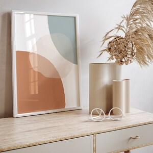 May include: A white framed print with an abstract design featuring muted tones of brown, beige, and blue. The print is displayed on a wooden desk with two drawers and a pair of white round glasses. A vase with dried flowers sits on the desk.