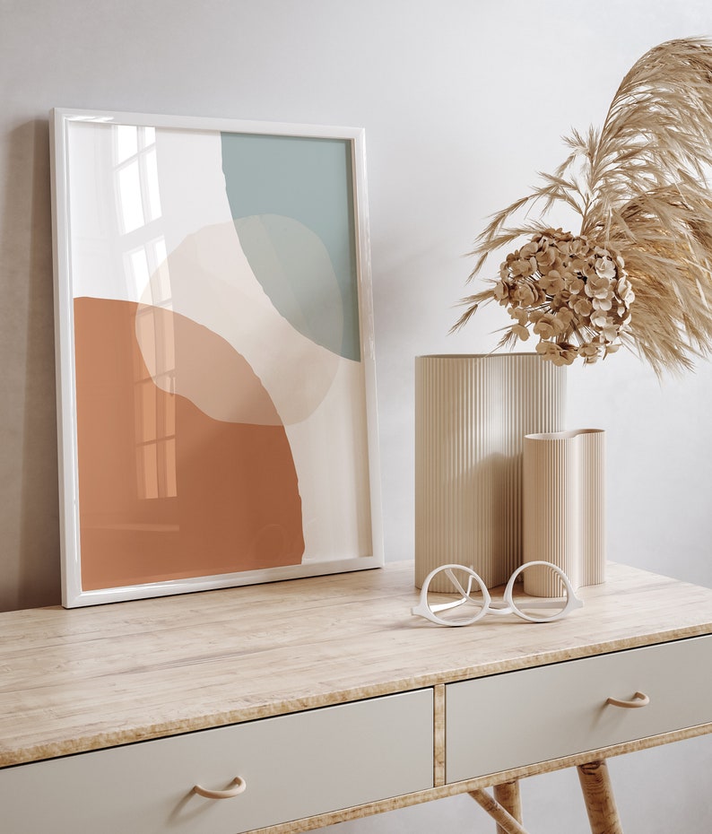 May include: A white framed print with an abstract design featuring muted tones of brown, beige, and blue. The print is displayed on a wooden desk with two drawers and a pair of white round glasses. A vase with dried flowers sits on the desk.