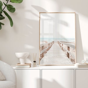 May include: A framed photograph of a wooden walkway leading to the sea. The walkway is made of light brown wood and has railings on either side. The sea is a light blue colour and the sky is a light grey colour. The photograph is in a light brown frame.