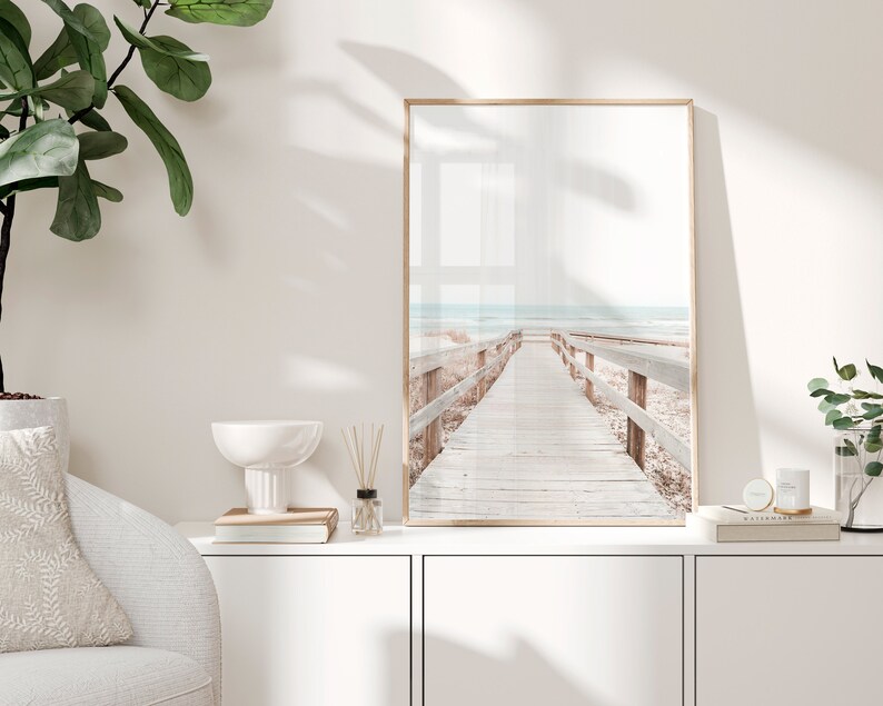 May include: A framed photograph of a wooden walkway leading to the sea. The walkway is made of light brown wood and has railings on either side. The sea is a light blue colour and the sky is a light grey colour. The photograph is in a light brown frame.