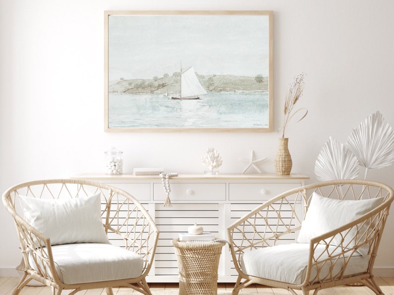 Puede incluir: Una pintura de acuarela de un velero en una masa de agua con un cielo azul claro y colinas verdes en el fondo. La pintura est&aacute; enmarcada en un marco de madera claro y est&aacute; colgada en una pared blanca. Dos sillas de mimbre con cojines blancos est&aacute;n frente a la pintura.