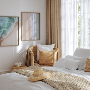May include: A bedroom with a white bed, a wicker chair, and two framed beach photos. The bed has a white duvet, a beige knitted throw, and a straw hat. The chair has a white pillow and a beige pillow. The room has a white wall and a window with white curtains.