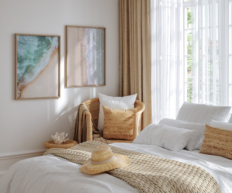 May include: A bedroom with a white bed, a wicker chair, and two framed beach photos. The bed has a white duvet, a beige knitted throw, and a straw hat. The chair has a white pillow and a beige pillow. The room has a white wall and a window with white curtains.