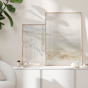 May include: Two framed photographs of ocean waves, one larger than the other, hanging on a white wall above a white cabinet. The cabinet has two doors and a countertop with a small bottle of diffuser sticks, a book, and a white candle.