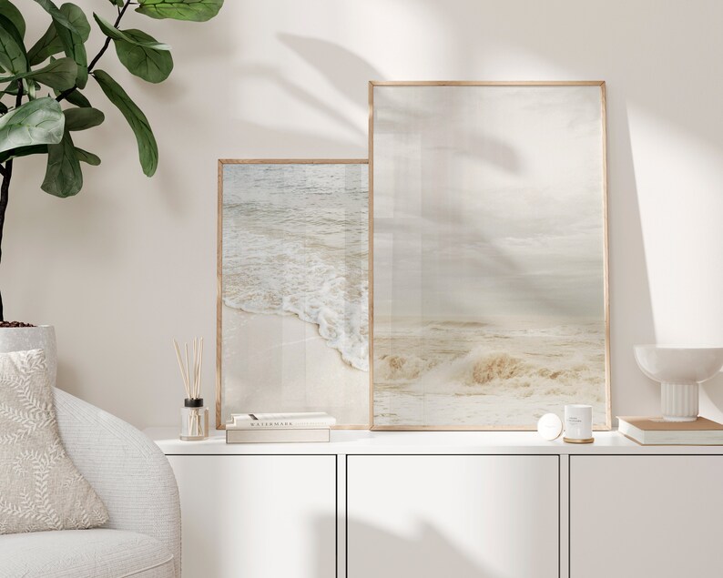 May include: Two framed photographs of ocean waves, one larger than the other, hanging on a white wall above a white cabinet. The cabinet has two doors and a countertop with a small bottle of diffuser sticks, a book, and a white candle.