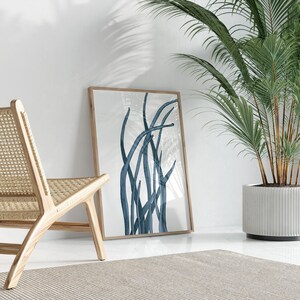 Op de afbeelding: Een ingelijste print van een abstract blauw ontwerp van gestileerde grassen of riet tegen een witte achtergrond. De print hangt aan een witte muur naast een houten stoel en een plant in een pot.