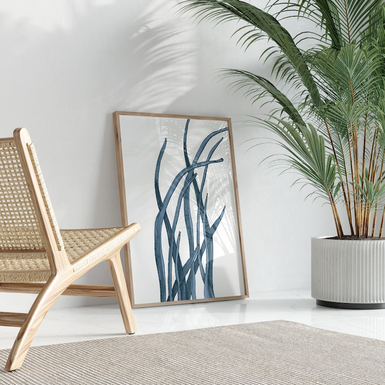 Op de afbeelding: Een ingelijste print van een abstract blauw ontwerp van gestileerde grassen of riet tegen een witte achtergrond. De print hangt aan een witte muur naast een houten stoel en een plant in een pot.