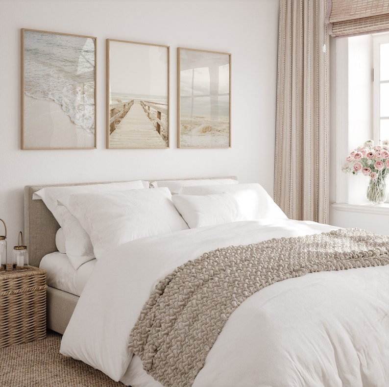 May include: A white bed with a beige knitted throw blanket and three framed beach-themed prints on the wall behind it. The bed has a beige headboard and white linens. The prints feature a beach scene with waves, a wooden walkway, and a beach with sand dunes.