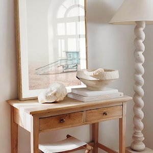 May include: A wooden table with two drawers and a white stool. On the table is a white sculpture, a book titled "Vincent van Gogh", and a white bowl. A framed photograph of a beach scene with a lifeguard stand is hanging on the wall behind the table.