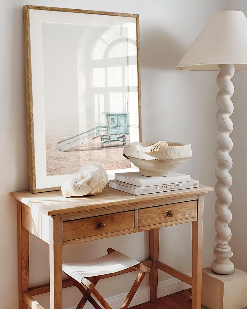 May include: A wooden table with two drawers and a white stool. On the table is a white sculpture, a book titled "Vincent van Gogh", and a white bowl. A framed photograph of a beach scene with a lifeguard stand is hanging on the wall behind the table.