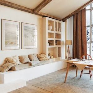 May include: A living room with a white couch, a wooden table, and a large window with a view of a snowy forest. The room is decorated with a brown rug, a lamp, and two framed pictures of seascapes.