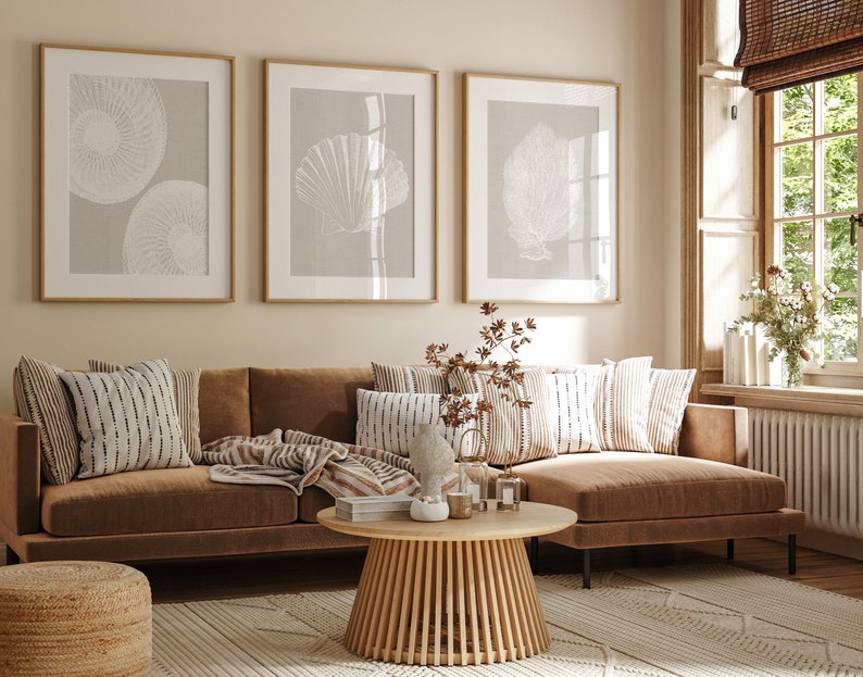 K&ouml;nnte beinhalten: Ein braunes Ecksofa mit einer Wolldecke und Kissen in einem Wohnzimmer mit drei gerahmten Drucken von wei&szlig;en Muscheln an einer beigefarbenen Wand.