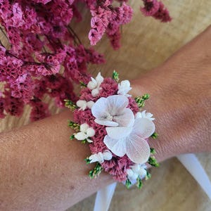 May include: A floral wrist corsage featuring deep magenta statice flowers, small white berries, and two large white hydrangea petals. The arrangement is secured with a white ribbon tie, creating a compact botanical accessory for the wrist.