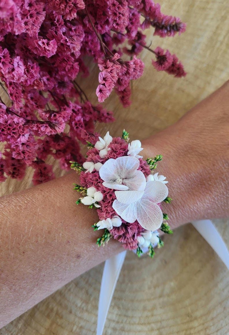 May include: A floral wrist corsage featuring deep magenta statice flowers, small white berries, and two large white hydrangea petals. The arrangement is secured with a white ribbon tie, creating a compact botanical accessory for the wrist.