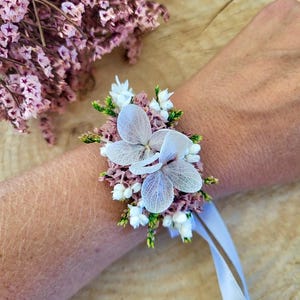 May include: A floral wrist corsage featuring two large, translucent white hydrangea petals centered among clusters of small, dried mauve statice flowers, tiny white blossoms, and green foliage, secured with a light blue satin ribbon tie.