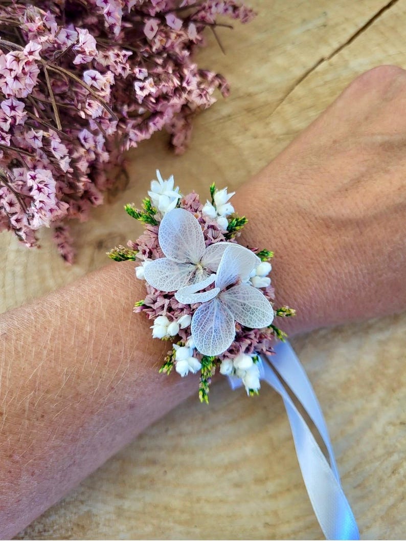 May include: A floral wrist corsage featuring two large, translucent white hydrangea petals centered among clusters of small, dried mauve statice flowers, tiny white blossoms, and green foliage, secured with a light blue satin ribbon tie.