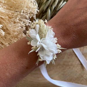 Peut inclure: Un bracelet de corsage composé de fleurs et de verdure séchées blanches et crème. L'arrangement floral est fixé avec un ruban blanc. Un accessoire délicat pour les mariages ou les événements spéciaux.